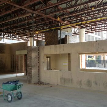 Interior of a warehouse with exposed ceilings, walls, cement flooring and a green wheel barrel.