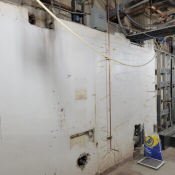 A construction site with a white wall and pipes running from the top down.